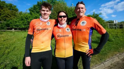 Deb Gascoyne The woman from the above picture, still wearing sunglasses but no helmet this time. On her left is a man and on her right is a teenage boy. All three have their arms around each other. They are standing in front of a grassy verge and wearing black lycra leggnigs and orange lycra tops reading: "Myeloma UK".