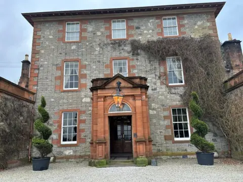 A large three storey sandstone building. There is gravel to the front and topiary on either side of a grand front door. 