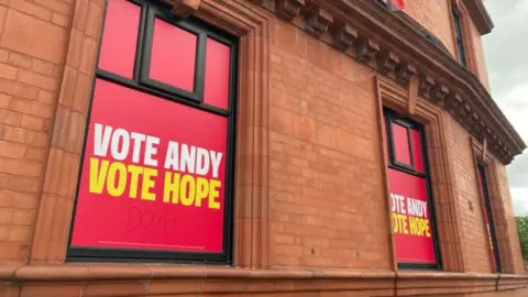 Jade Taylorson/BBC A large, Victorian style red-brick building with the windows covered in red posters with the slogan "Vote Andy, Vote Hope" in white and yellow lettering. 