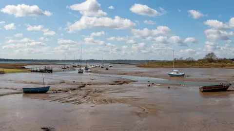 Suffolk County Council A bay with the tide out and boats on the sandbanks. It is a sunny day with white clouds in the sky.