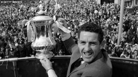 Black and white image of Dave MacKay, facing the camera, holding aloft the FA Cup over a handrail.  Below, a crowd of fans.