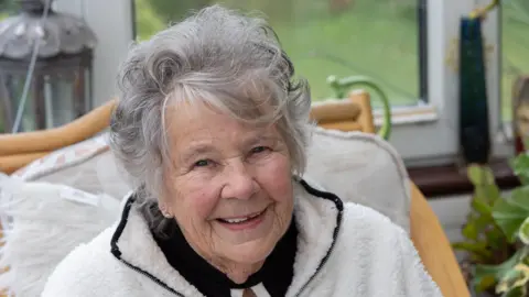 Chris George A close up shot of a smiling woman with greying hair sitting in front of a window