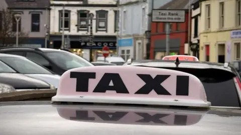 The top of a car is visible with a large black and white taxi sign on top. The sign can be seen reflected on the roof of the vehicle. There are other parked cars and a colourful mix of buildings in the background.