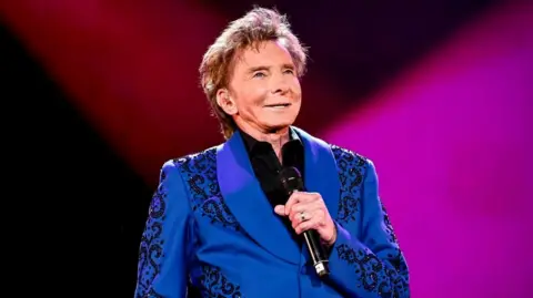 Barry Manilow wearing a blue suit with a black design, holding a microphone up
