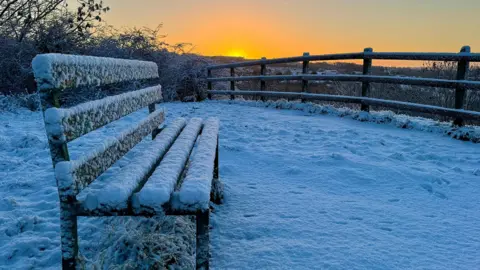 A park bench covered in snow overlooking a snowed over fence while there is an organe glow in the sky and the sunsets.