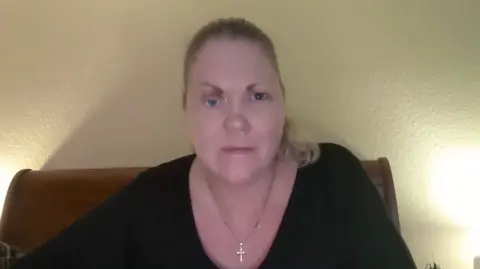 A woman sat on a bed smiling at her computer camera. She is wearing a black long sleeve top and wearing a cross necklace.