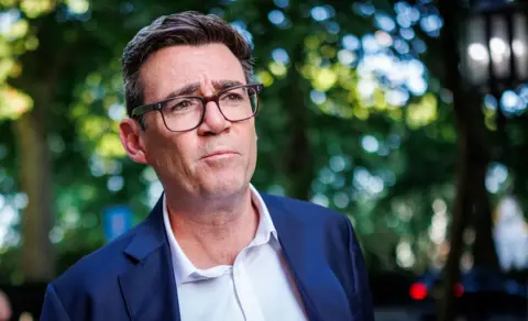 Dan Kitwood / Getty Images A head and shoulders crop of a photo of Andy Burnham wearing a suit and shirt, with black rimmed glasses against a bokeh background of green folioage, pictured in Millbank on 2 July.