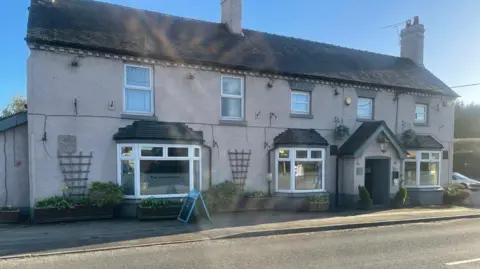 The pub stands on the side of a road with a wooden board outside. It is painted a light colour with windows on the top and bottom levels.