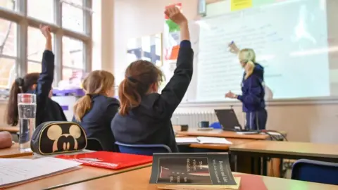 File photo of children in a classroom looking at a teacher standing by a whiteboard. Two have their hands up.