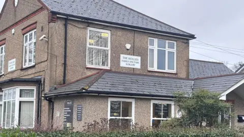 The front of The Hollies Surgery, which is a beige-coloured, two-storey pebble-dash house. A sign reads THE HOLLIES FAMILY DOCTORS SURGERY.