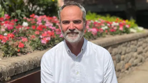 BBC A person in a white button-up shirt sits on a wooden bench beside a raised flower bed filled with pink, red and white flowers. Trees and a residential building are visible in the background on a sunny day.