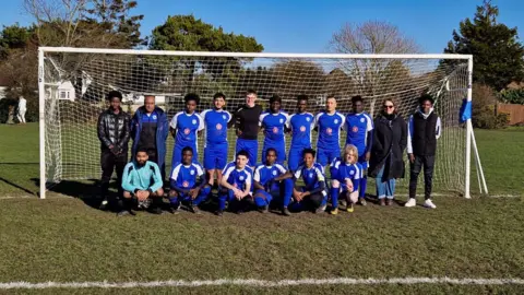Ellie Douglas The group of players wearing their blue and white football kits and stood around posing for a photo in front of a goal