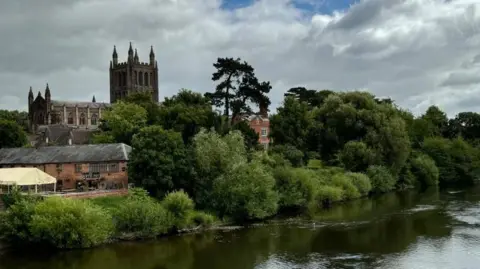 A flowing river with trees and bushes alongside, historical buildings and a cathedral in the background. 