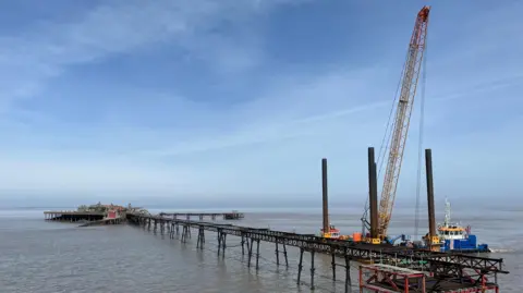 A wide shot of Birnbeck Pier taken from the land. The pier has a long metal walkway leading out to it, and two barge-type boats are moored alongside and a large crane is towering over the structure. In the distance the main island of the pier is visible.