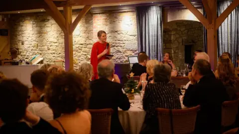 Somerville Captures Caitlin Riddell is standing at the front in front of people seated at round tables. She is holding a microphone and is wearing a red dress  