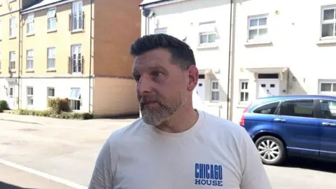A man with blue eyes and short brown/grey hair and a beard standing on Redhouse Way in Swindon, straight-faced and wearing a white t-shirt with terraced houses, apartments and a blue car in the background.