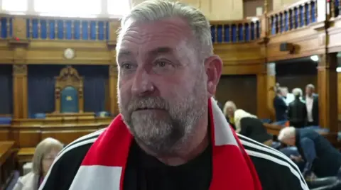 Perry McMillan, who has a grey beard and smartly combed grey hair, wears a red and white Southampton FC scarf inside a historical wood-panelled council chamber.