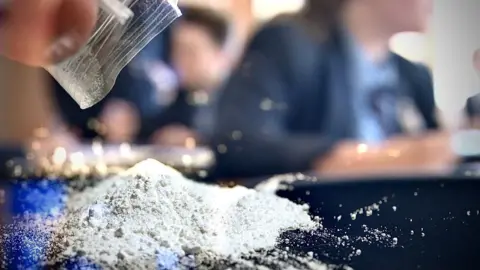 A pile of white powder has just been poured from small plastic bag on to a desk in a stylised image with young people in school uniform blurred in the background. 