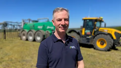 A white man with short white hair looks at the camera. He is wearing a blue polo shirt with the letters "NFU" on it. In the background is a yellow tractor with a green tank attached to the back of it.