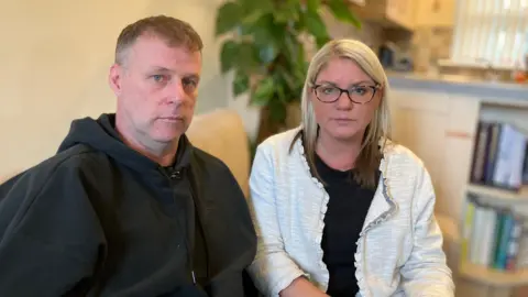 Ben's parents are sat inside a house with a kitchen, bookshelves and plant in the background. They are both straight-faced, looking at the camera.