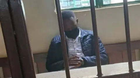 Festus Arasa Omwamba sits behind bars with his hands clasped, wearing a black face mask