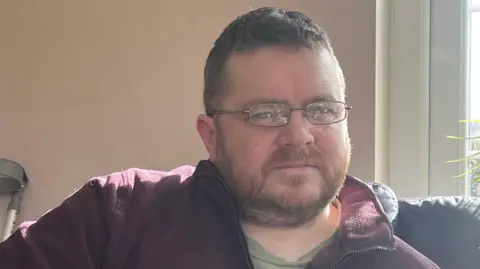 Mark has thin rectangular glasses and is wearing a burgandy fleece unzipped at the collar. He has short stubble and cropped brown hair.