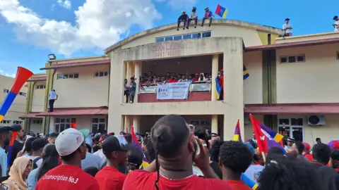 BBC/Yasine Mohabuth Dozens of people stand in the streets of Port Louis. They wave the red, blue, yellow, and green flag of Mauritius. A few people sit on top of a yellow building, looking down at the large crowd. 