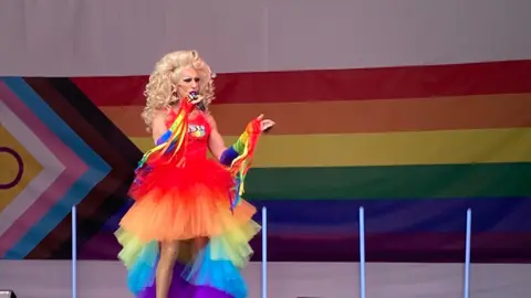 A drag artist on stage wearing a multi-coloured rainbow chiffon dress. She has big hair with lots of blonde curls. Behind her is the LGBTQIA+ flag.