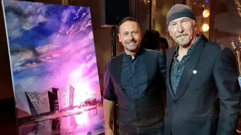 Two men are standing next to each other. A painting of the Titanic building is next to them. Both men are wearing suits.
