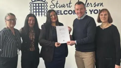 Stuart Anderson A man smiles as he presents a woman with a certificate. Two other women are standing to her left and another woman is to the man's right.