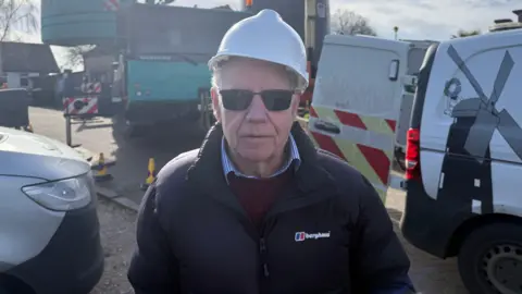 Grace McGrory/BBC A man wearing a black coat, a brown jumper and white shirt is wearing white helmet and black sunglasses. He is stood in front of two vans and a green and black crane can be seen behind him.
