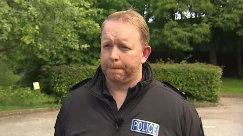 A picture of Andy Wills looking to the left side of the camera. He is wearing a black jacket with a police badge on it. He has short hair and a short beard. He is stood in front of a hedge and trees.