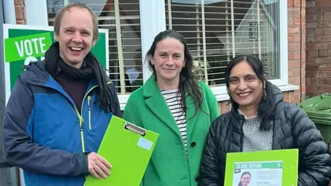 A man on the left (Jonathan Chilvers) smiling in a bluejacket holding a green clipboard next to Clare Bryan who smiles in a green jacket. On her left is Hema YellaPragada who is also smiling holding a green clipboard, she wears a black jacket.