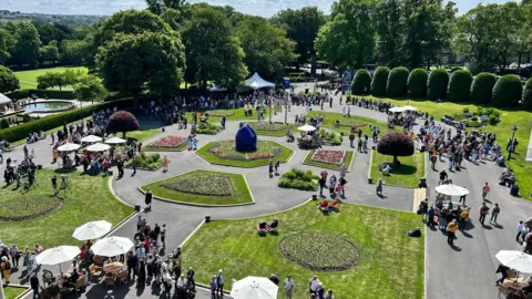 An aerial view of Lister Park in Bradford, a structured garden with flower beds, trees and bushes and lots of people milling about with parasols dotted around. 