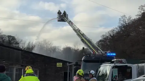 BBC A large platform extended from a fire engine is holding two fire and rescue crew members, with water from a hose come down to the left onto a roof, and the air is smoky. You can see in the foreground three crew members with helmets and in high vis jackets. There are trees all around.