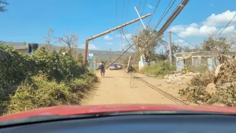 supplied A driver's viewpoint from within the car shows a large wooden pole with wires bent over a dusty road on a sunny day