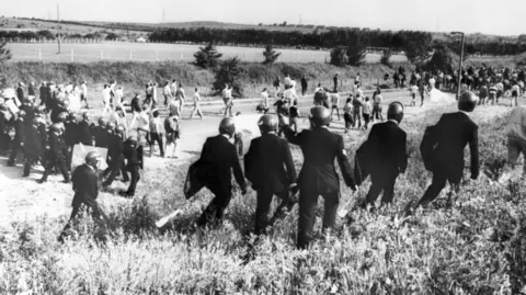 A black and white photo shows police in uniform with helmets and riot shields in a green field with miners in jeans and t-shirts