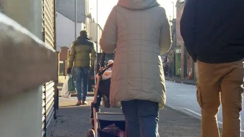 People photographed from behind walking down a street in Gloucester. A couple are seen in coats walking with a pram, and further ahead a man in a coat and gloves carries shopping bags. 