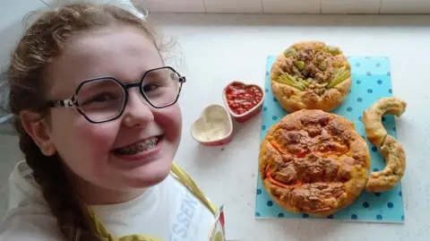 Lyla has long brown hair and her hair is in pigtails. She has round colourful glasses and is smiling. She is standing beside a number of delicious-looking baked goods and sauces. 