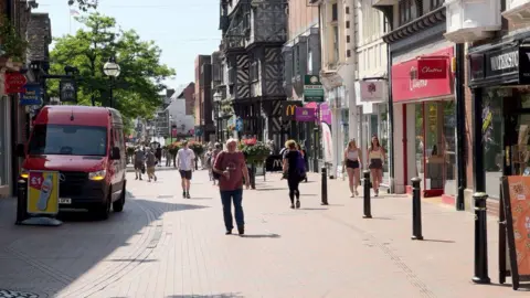 A busy town centre, with a view along a pedestrianised street. There is a red van parked on the left hand side of the image facing the camera. On the right hand side are a number of shops and partway down the street is a black and white timber-framed building.