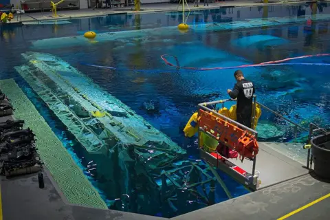 Kevin Church/BBC Wide of support team member standing on a platform overlooking the water of the pool. Some of the modules of the space station are clearly visible below the surface. There is a row of scuba equipment to the left of the picture and there are red and blue " umbilical hoses " stretching down to the unseen astronauts beneath the water's surface. 