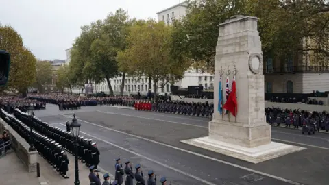 A remembrance ceremony at The Cenotaph in Whitehall, London. Flags of the United Kingdom, Royal Air Force, and Red Ensign are prominently displayed and uniformed military personnel are lined up in a formal formation. Members of the public and officials observing the event from behind barriers.