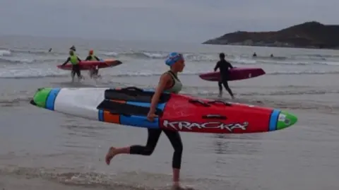 A woman in a wetsuit holding a surf board running along a beach. The sky is grey. There are people running into the sea in the background. 