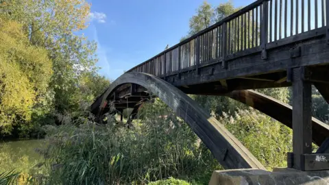 A wooden footbridge spanning a river. The bridge is supported by arches. There is vegetation in the foreground and trees in the background along the banks of the river.