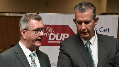 Two men, both with grey hair, wearing grey suits with blue ties. They are stood in front of a DUP banner and engaged in conversation. On the left, Jeffrey Donaldson is wearing black rimmed glasses and appears to be speaking.