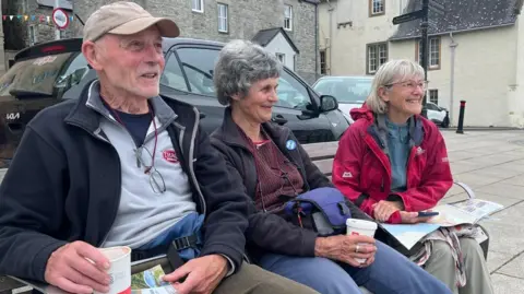 Ruth Nutman sits between two friends on a bench. Joy as short grey hair and she is wearing a black fleece over a burgundy top.