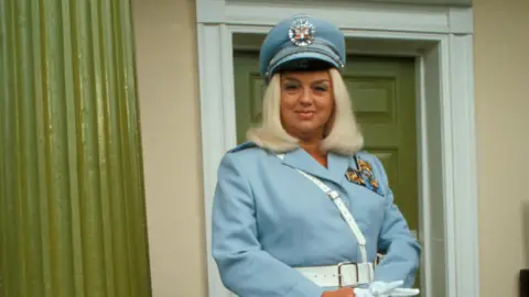 Diana Dors in a blue and white uniform, including a hat, and holding white gloves.