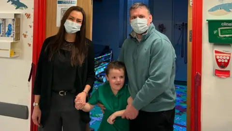 Gemma and Robert standing with Ellis outside the immersive room. Gemma, on the left, wears a black blouse, black blazer and grey trousers. She has long dark brown hair and is wearing a blue hospital mask. Ellis, in the middle, wears a green t-shirt and has short blonde hair. Robert, on the right, wears a blue quarter zip sweatshirt and a mask.