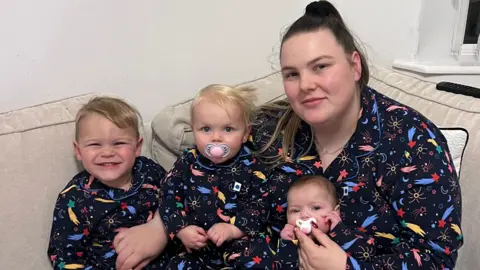 Paige Carr, in her late 20s, with brown hair tied back into a ponytail, sits on a cream couch holding a newborn baby, a toddler and a little boy on her knee. They are all wearing matching dark blue pyjamas with a shooting star design. 