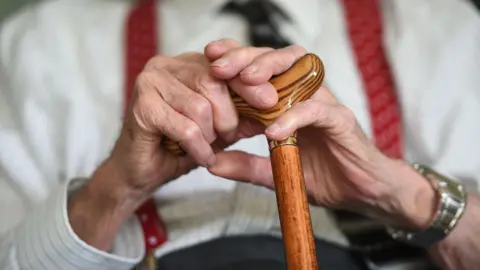 An elderly man with red braces holds his walking stick
